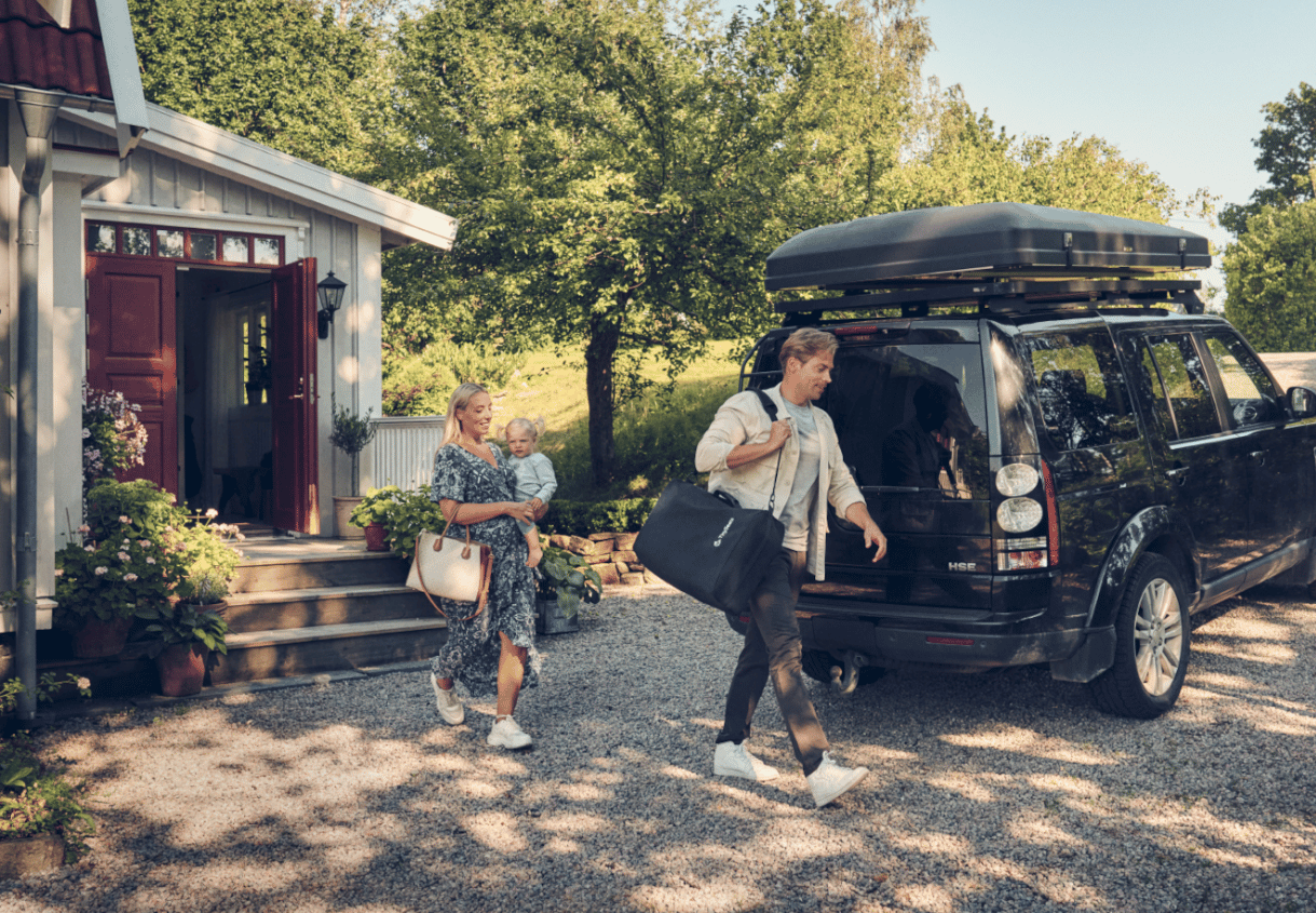 Its summer in Sweden and two people are walking towards a black SUV, with man carrying a portable extended rear-facing car seat in a shoulder bag. 