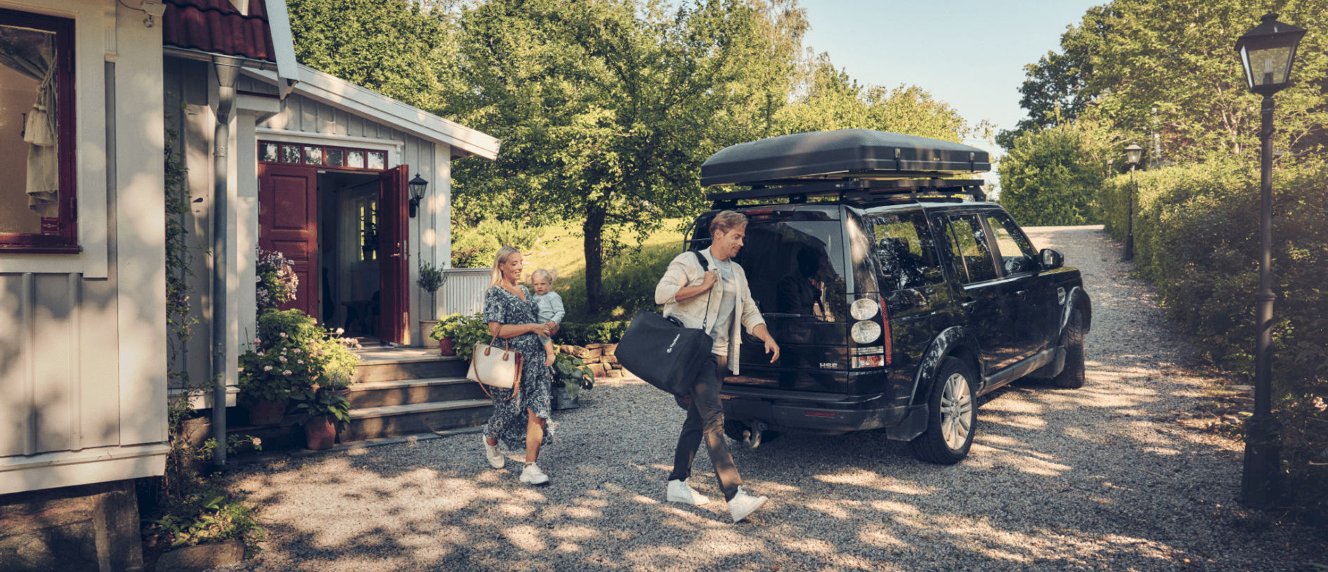 Its summer in Sweden and two people are walking towards a black SUV, with man carrying a portable extended rear-facing car seat in a shoulder bag. 
