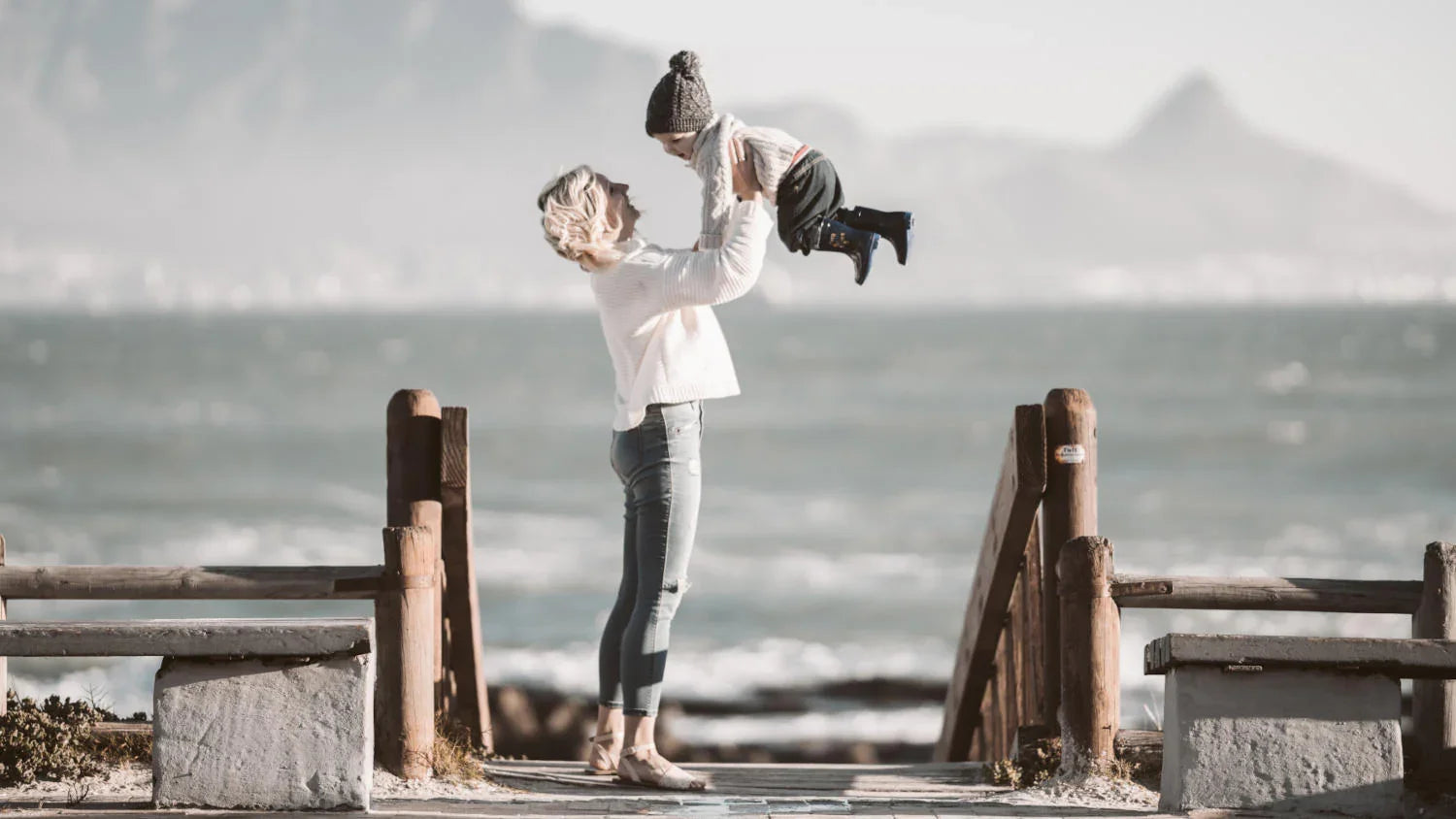 A mother holds up her son in her arms on a windy beach during an autumn morning in Sweden.